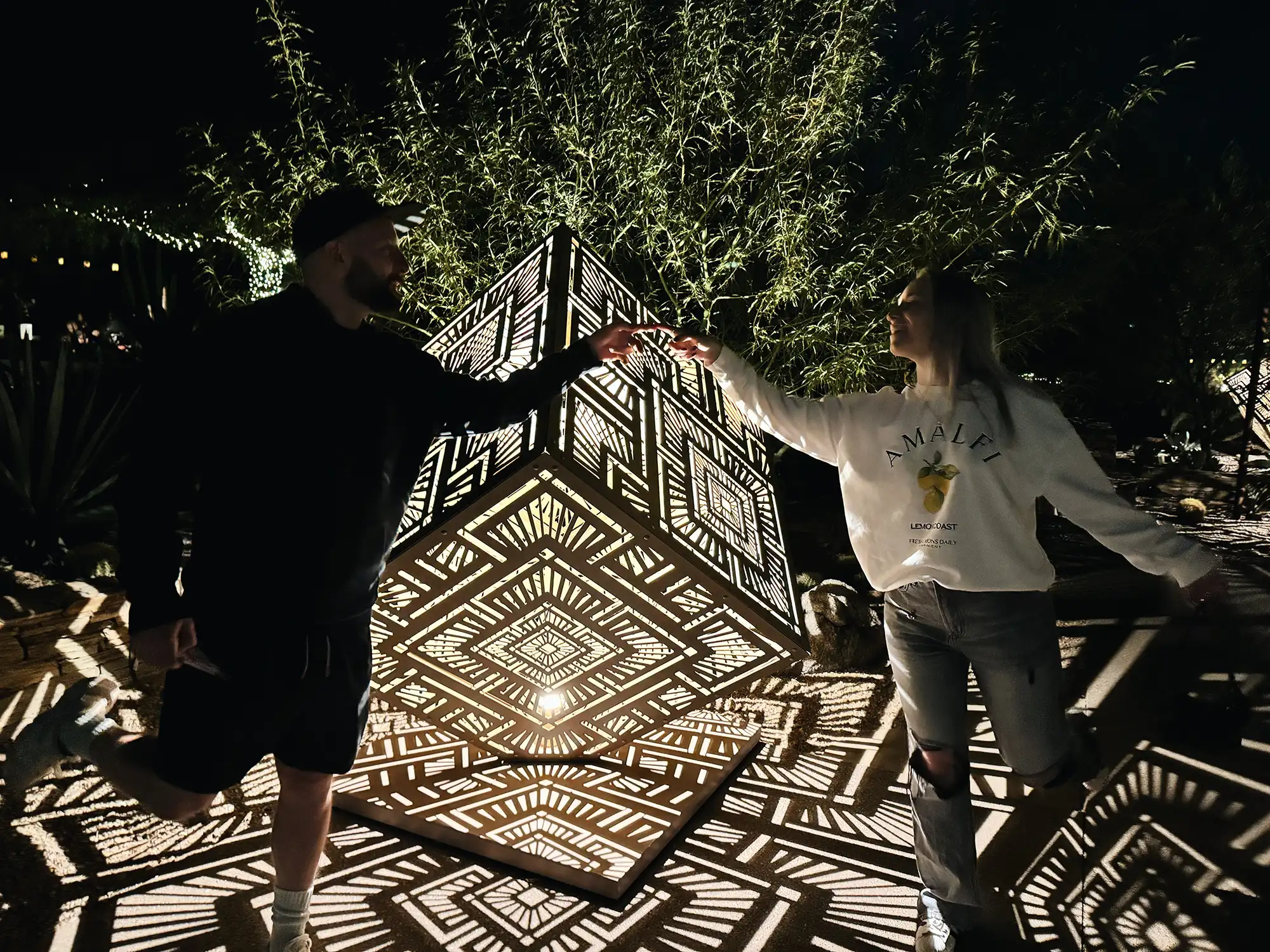 alt: Two people in front of a geometric light sculpture posing, reaching out to each other and touching fingers