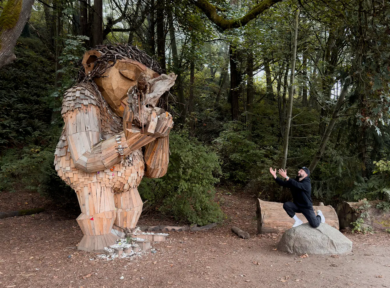 alt: A man kneeling in front of a large wooden troll statue in a forest.